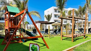 A playground with swings and a slide, surrounded by palm trees and a pool area. - Almarena Apartments Coto Coral (Mazatlán)