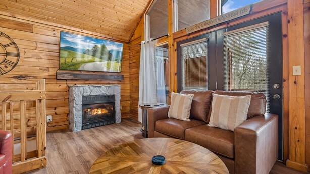 Un intérieur de chalet chaleureux avec une cheminée, un canapé en cuir, un plancher en bois et une grande fenêtre avec des stores.