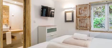 A hotel room with a wooden door, a television mounted on the wall, a bedside table, a bed with two pillows, and a window with a view of greenery.
