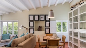 Ein gemütliches Wohnzimmer mit einer Couch, einem Esstisch und Stühlen. An der Wand befinden sich gerahmte Bilder, eine Hängeleuchte und ein Schrank mit Glastüren.