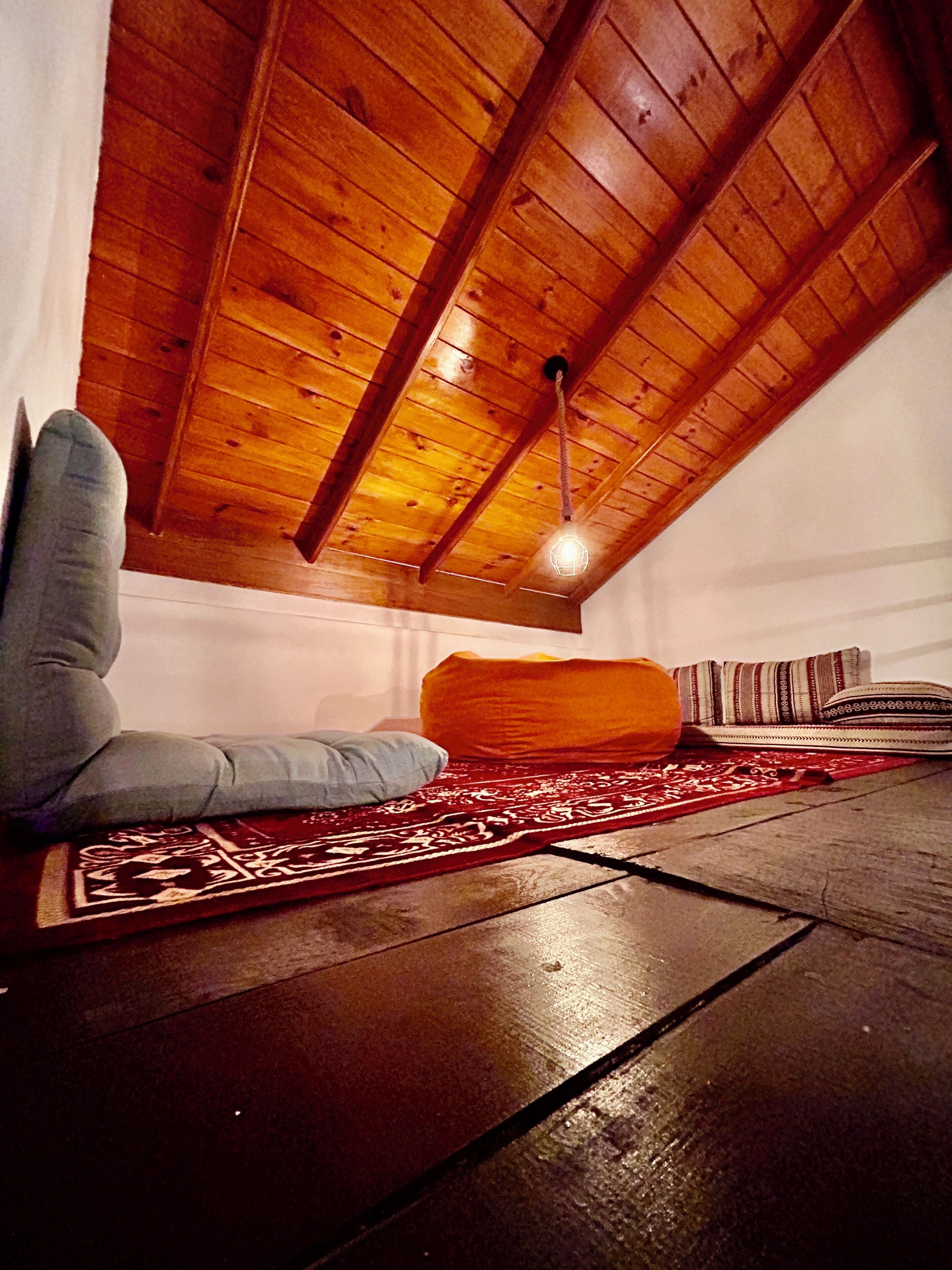 A cozy loft bedroom with a sloped ceiling, a hanging light fixture, a patterned rug, and a large orange cushion.