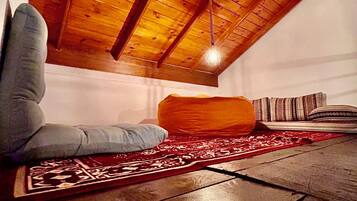 A cozy loft bedroom with a sloped ceiling, a hanging light fixture, a patterned rug, and a large orange cushion.