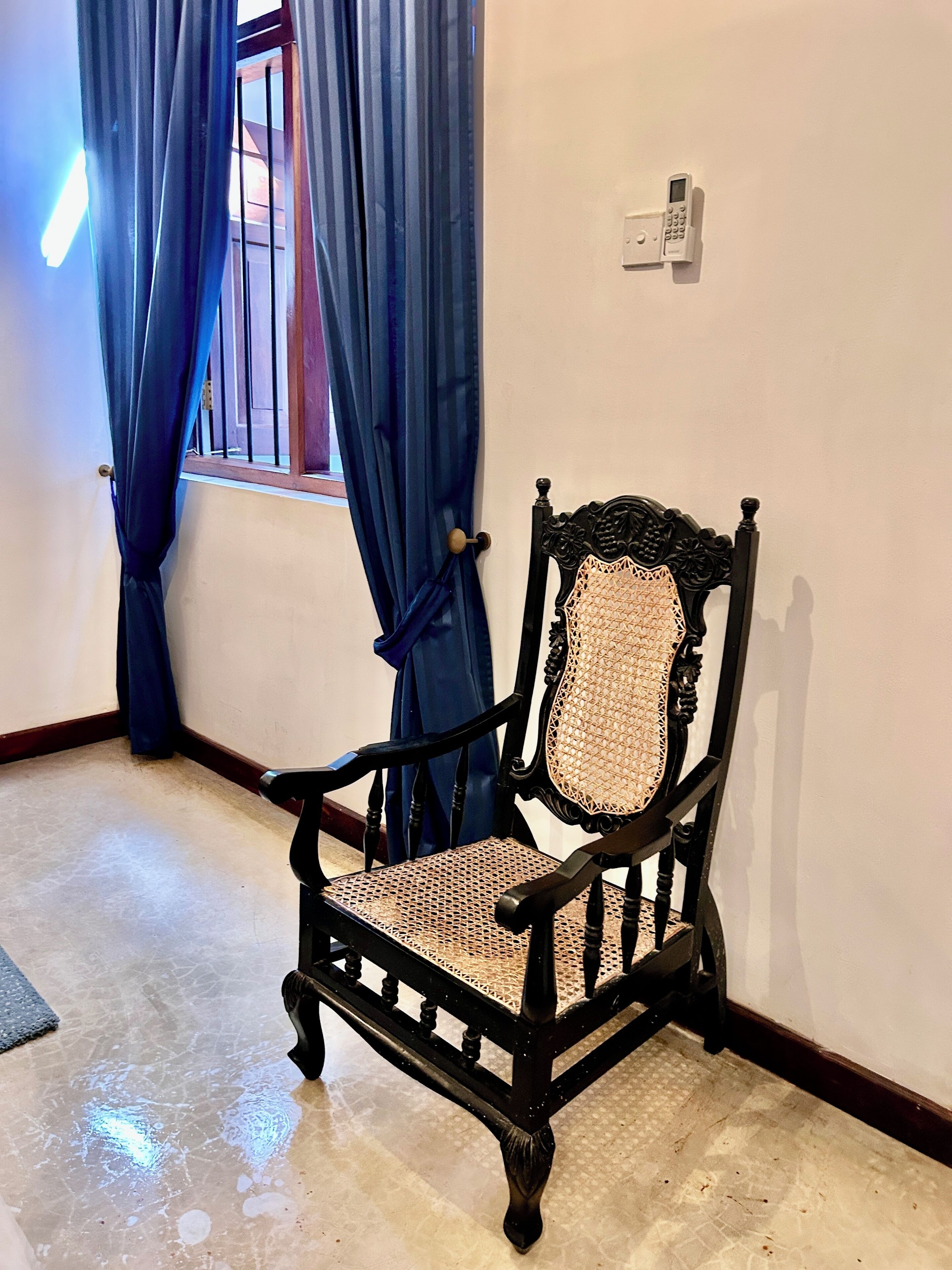 A corner room with a wooden chair with a woven seat and backrest, a window with blue curtains, and a wall-mounted light switch.