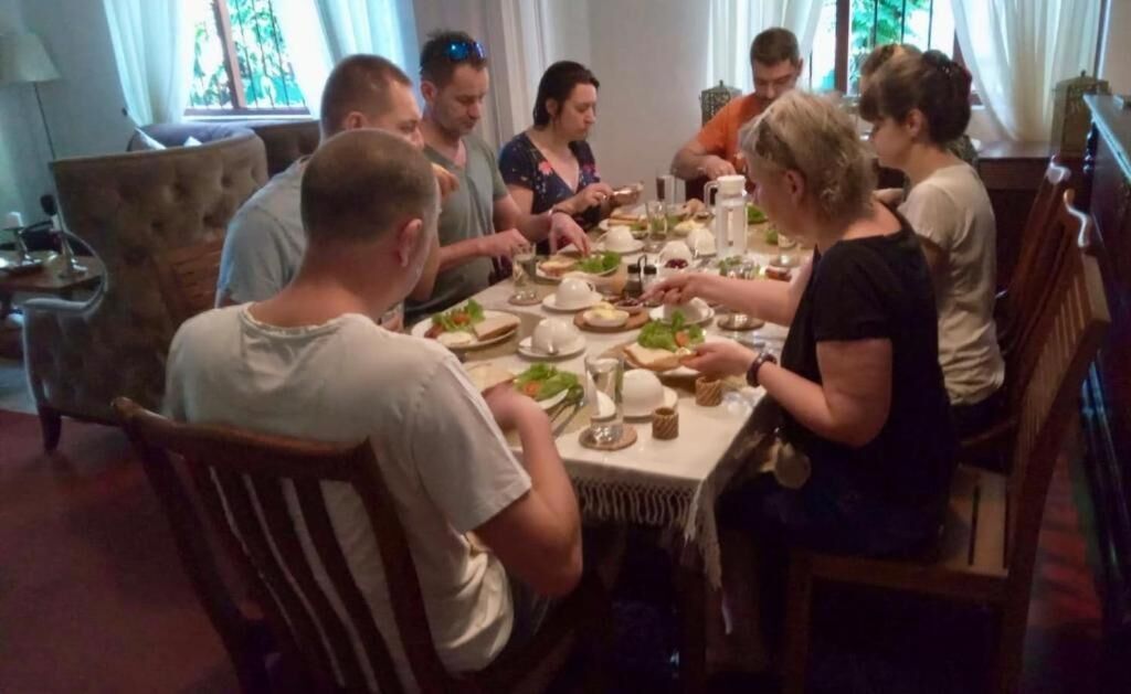 A group of people seated around a dining table in a room with a chandelier and a window with curtains.