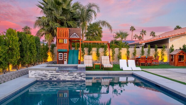 A swimming pool with lounge chairs, a wooden playhouse, and a palm tree.