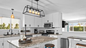 A modern kitchen with white cabinets, granite countertops, a built-in oven, and a coffee maker. There are two stools and a small dining table.