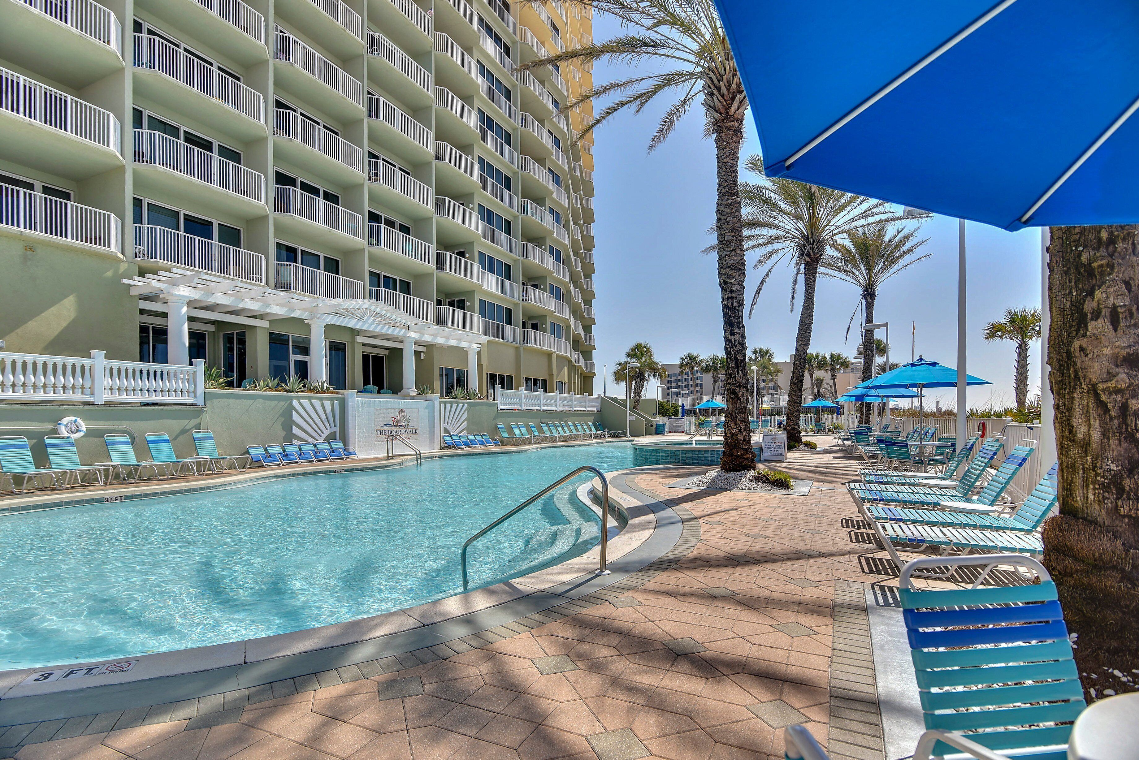 Área de piscina de hotel, com uma piscina grande, espreguiçadeiras e um edifício com várias andares e varandas.