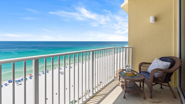 Ein Balkon mit Korbstuhl und Tisch, der Blick auf einen Strand mit Sonnenschirmen und Menschen bietet.