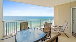 A balcony with a glass table and wicker chairs, overlooking a beach with clear blue water and a distant shoreline.