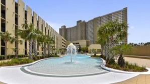 A swimming pool with a central fountain, surrounded by palm trees and modern buildings.