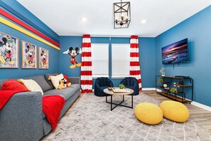 A living room with a blue wall, Mickey Mouse mural, red and white curtains, a grey sofa, two armchairs, a coffee table, a TV, and a bookshelf.