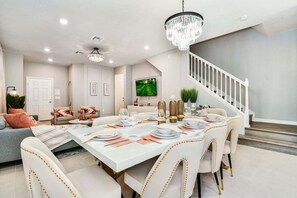A dining area with a white table set for six, a chandelier, and a staircase leading to another level.
