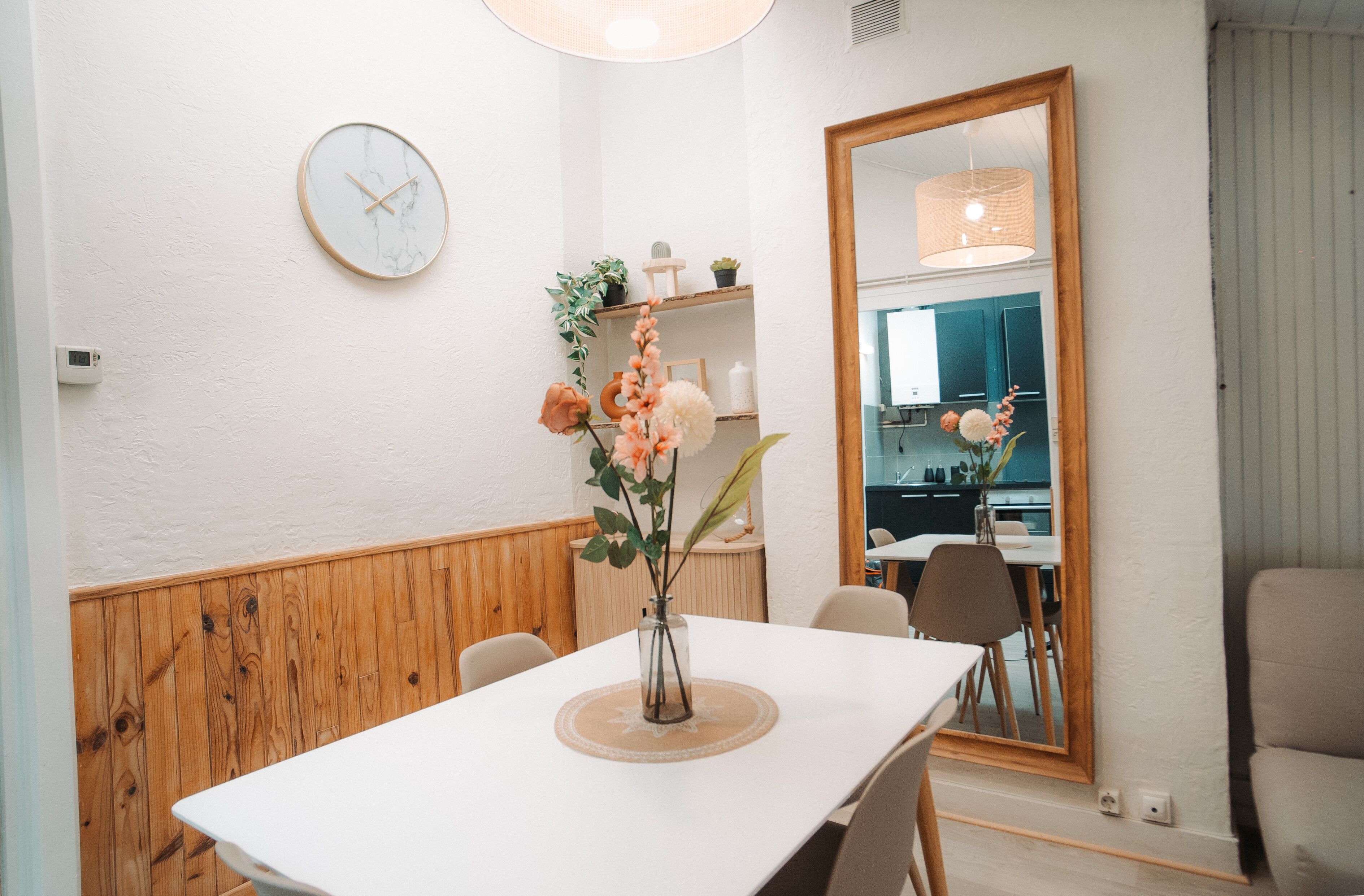 A dining area with a white table, chairs, a vase with flowers, a clock, and a mirror.
