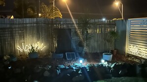 Un pati nocturn amb tanca de fusta, plantes en testos, una barbacoa i una làmpada amb energia solar.
