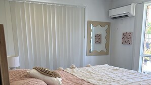 Un dormitori amb estructura de llit de fusta, un ventilador de sostre, parets blanques i una finestra amb persianes.