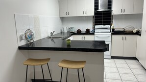 Una cuina moderna amb armaris blancs, un taulell negre i dos tamborets de fusta.