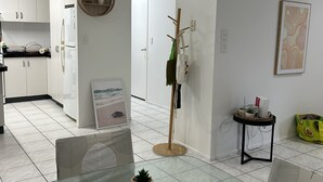 A modern living room with a glass coffee table, a dining area with chairs, a kitchen with white cabinets, and a hallway with a coat rack. - Beach, Sand Relax (Bucasia)