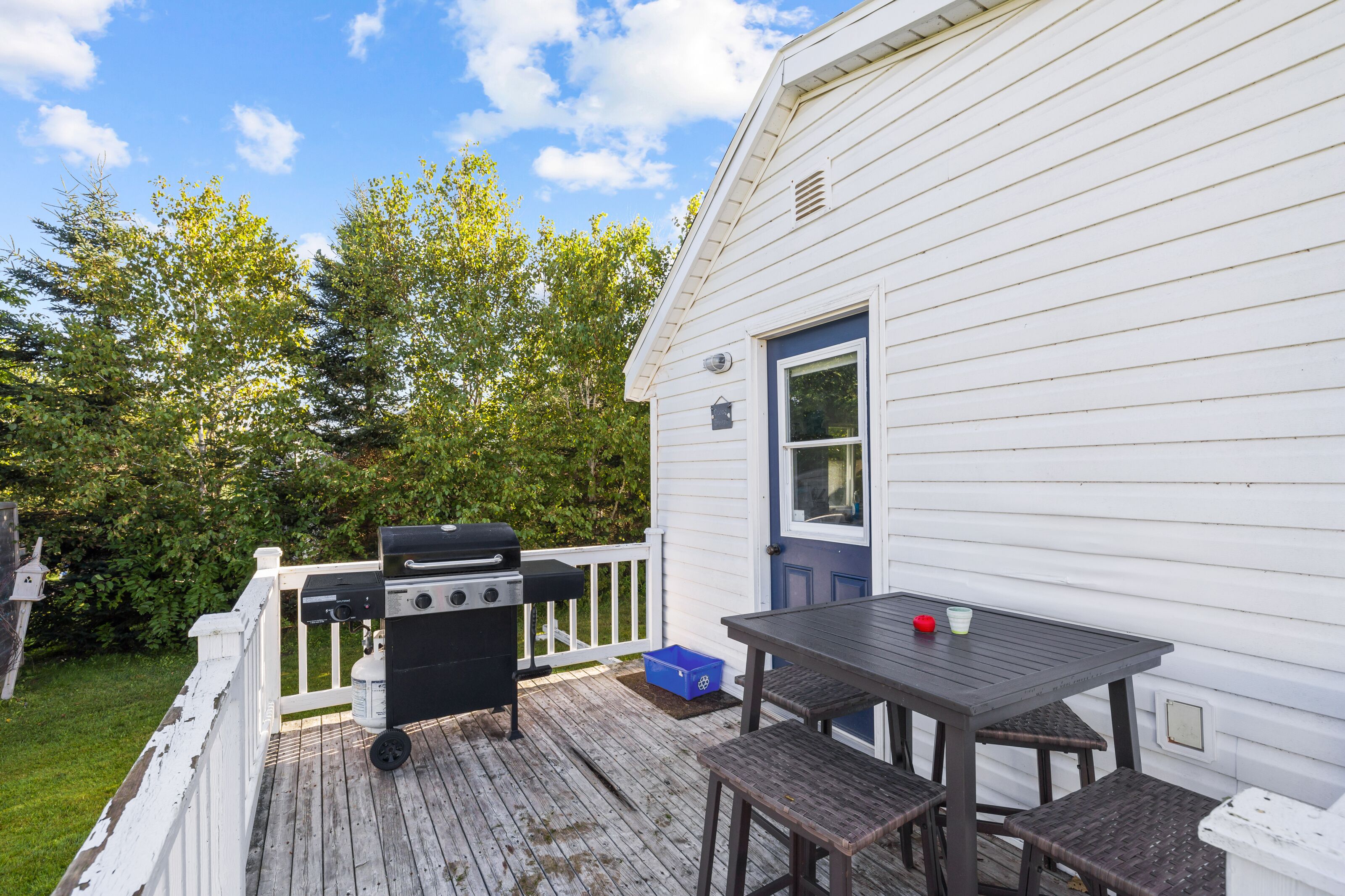 A deck with a grill, a table, and chairs, adjacent to a white house with a blue door.