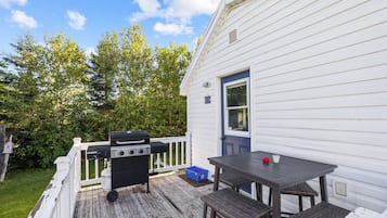 A deck with a grill, a table, and chairs, adjacent to a white house with a blue door.