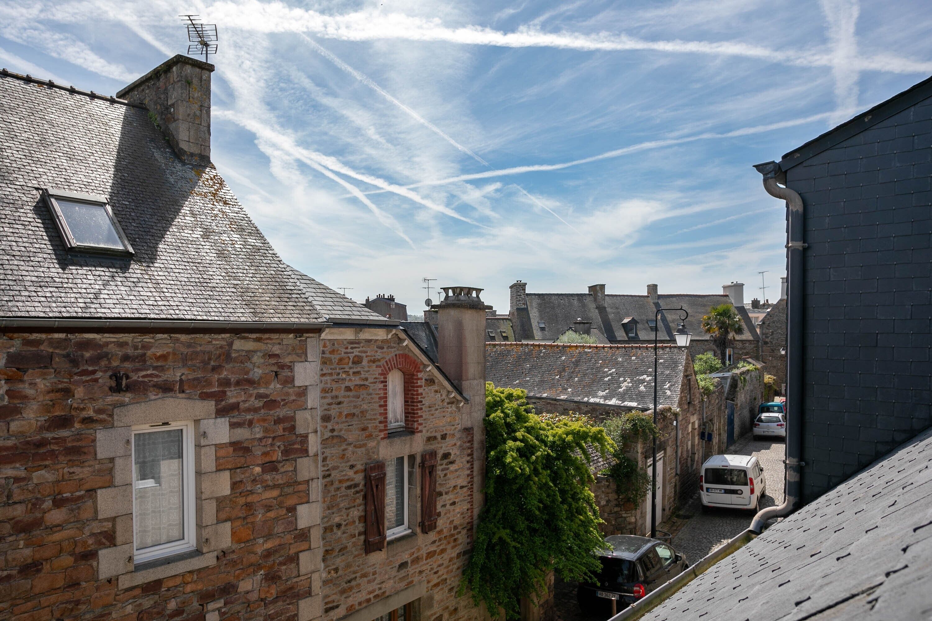 A narrow street with stone buildings, a car parked on the side, and a clear sky with contrails.