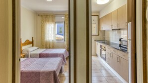Una habitación con dos camas, una ventana con cortinas, una kitchenette con armarios y electrodomésticos, y piso de baldosas.