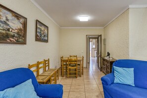 A living room with a blue sofa, a wooden dining table with chairs, and a hallway leading to another room. - Apartment 'La Siesta' with Private Terrace and Wi-Fi (L'Escala)