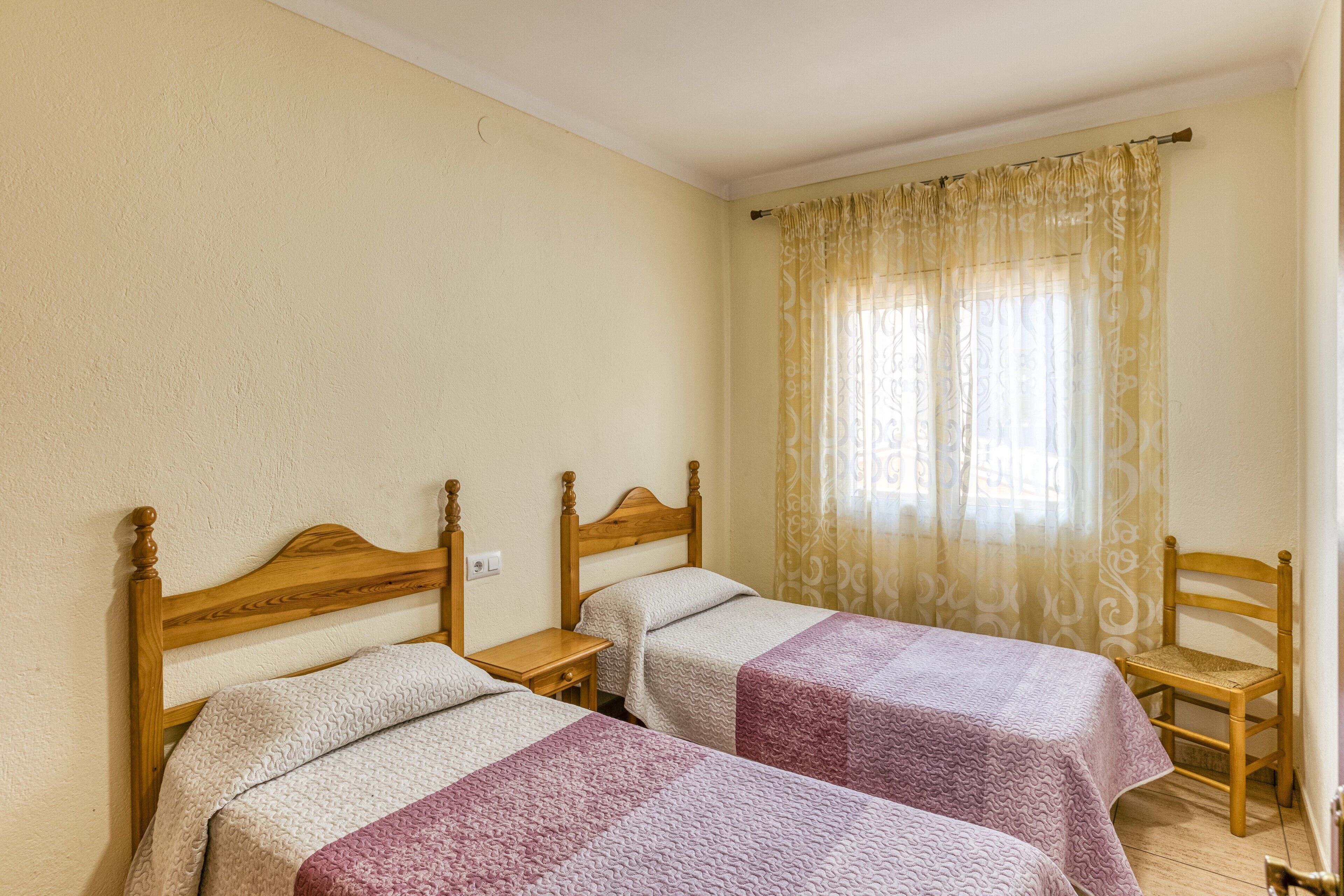 A room with two wooden beds, a wooden chair, and a window with patterned curtains.