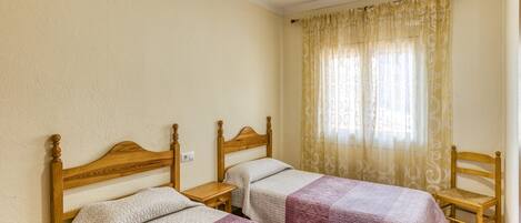 A room with two wooden beds, a wooden chair, and a window with patterned curtains.