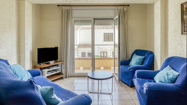 Una sala de estar con una televisión, dos sillones azules, una mesita y un ventanal corredera que da a un balcón.