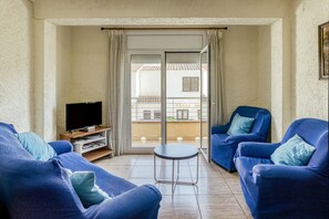 Ein Wohnzimmer mit Fernseher, zwei blauen Sesseln, einem kleinen Tisch und einer Schiebetür, die auf einen Balkon führt.