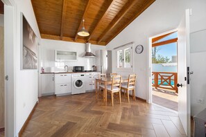Cozinha com armários brancos, máquina de lavar, mesa de jantar com cadeiras e vista para uma piscina através de uma porta aberta.