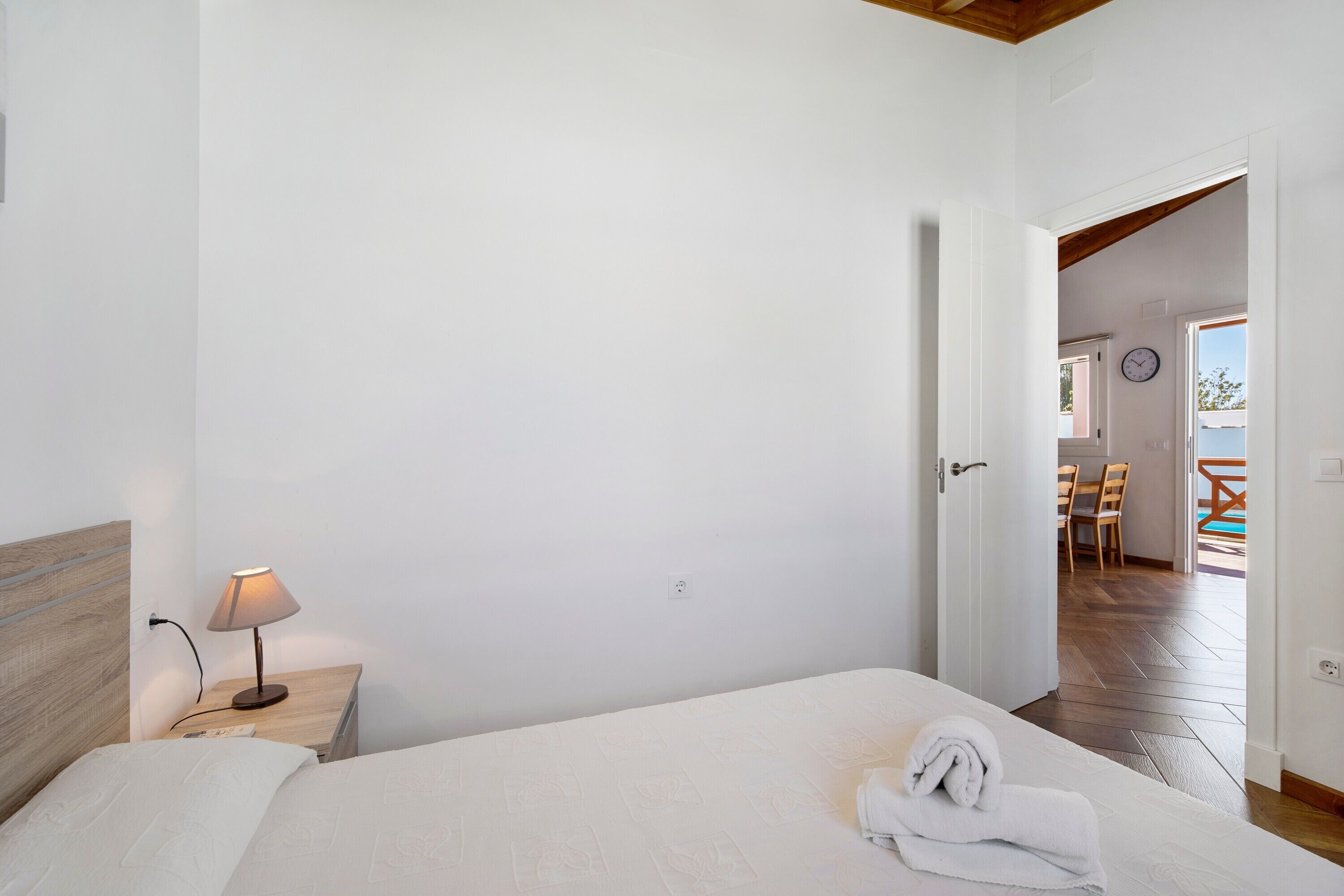 A bedroom with a bed, a bedside lamp, a wooden headboard, and a view of an outdoor patio with a table and chairs.