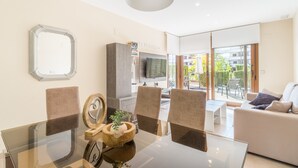 Ein modernes Wohnzimmer mit einem Glastisch, bequemen Sitzgelegenheiten, einem Flachbildfernseher und Blick ins Freie.
