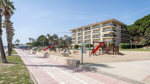 Ein Spielplatz direkt am Strand mit Schaukeln und Rutschen, einem Sandbereich und einem mehrstöckigen Gebäude im Hintergrund.