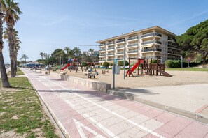 Un'area giochi sulla spiaggia con altalene e scivoli, una zona sabbiosa e un edificio a più piani sullo sfondo.