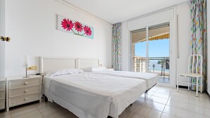 Ein Hotelzimmer mit zwei Betten, einem Nachttisch, einem Sessel und einem Balkon mit Aussicht. An der Wand hängt ein Gemälde.