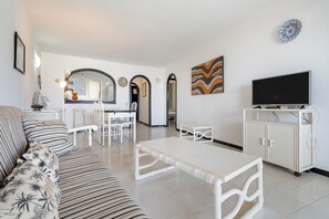 A living room with a sofa, a coffee table, a TV, and a cabinet. There is a dining area with chairs and a table in the background.