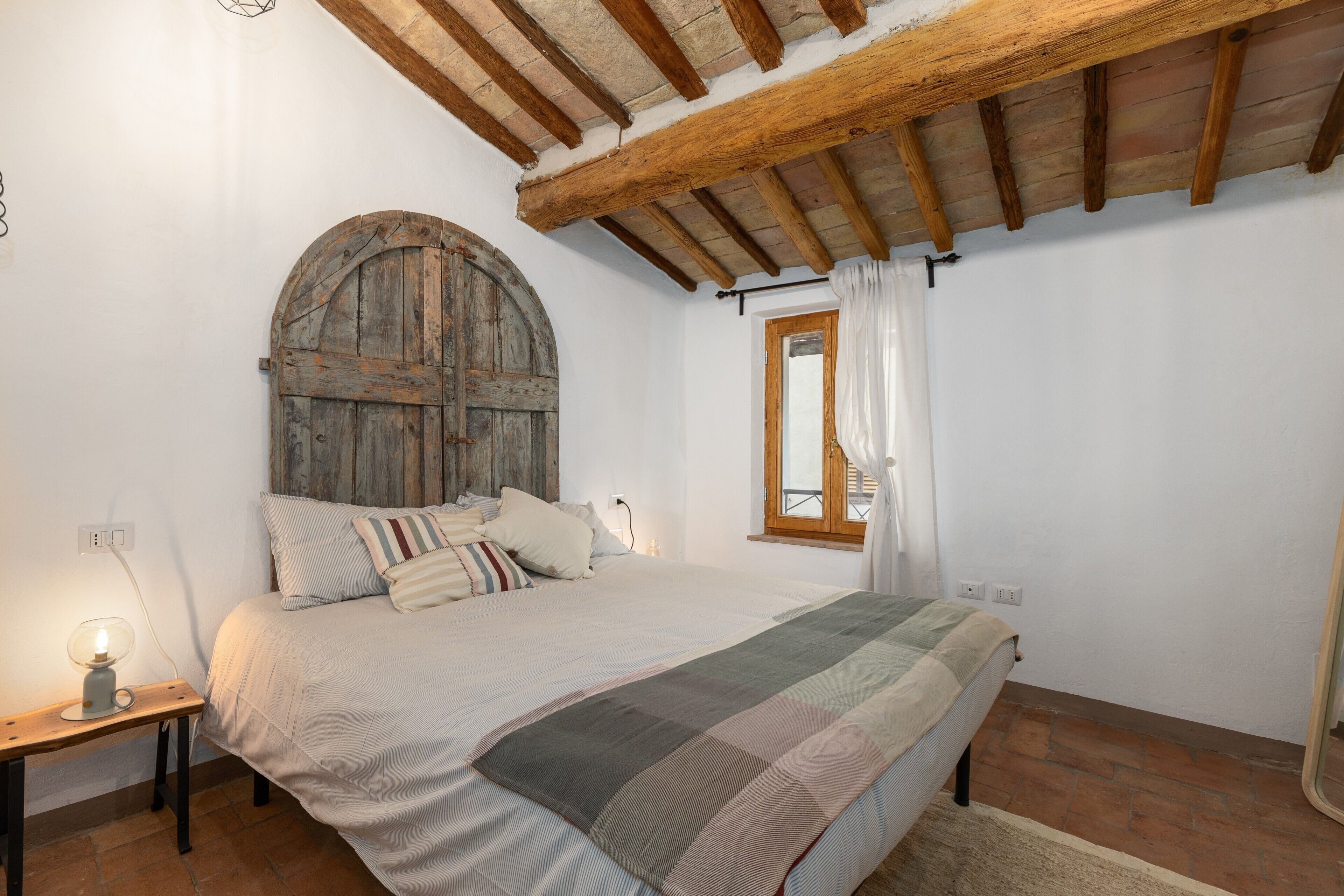Ein Schlafzimmer mit Holztür, einem Bett mit gestreifter Decke, einem Nachttisch mit Lampe und einem Fenster mit Vorhängen.