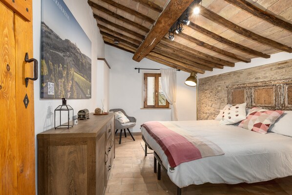 A bedroom with a wooden ceiling, a bed with a patterned bedspread, a desk with a lamp, a chair, and a stone wall.