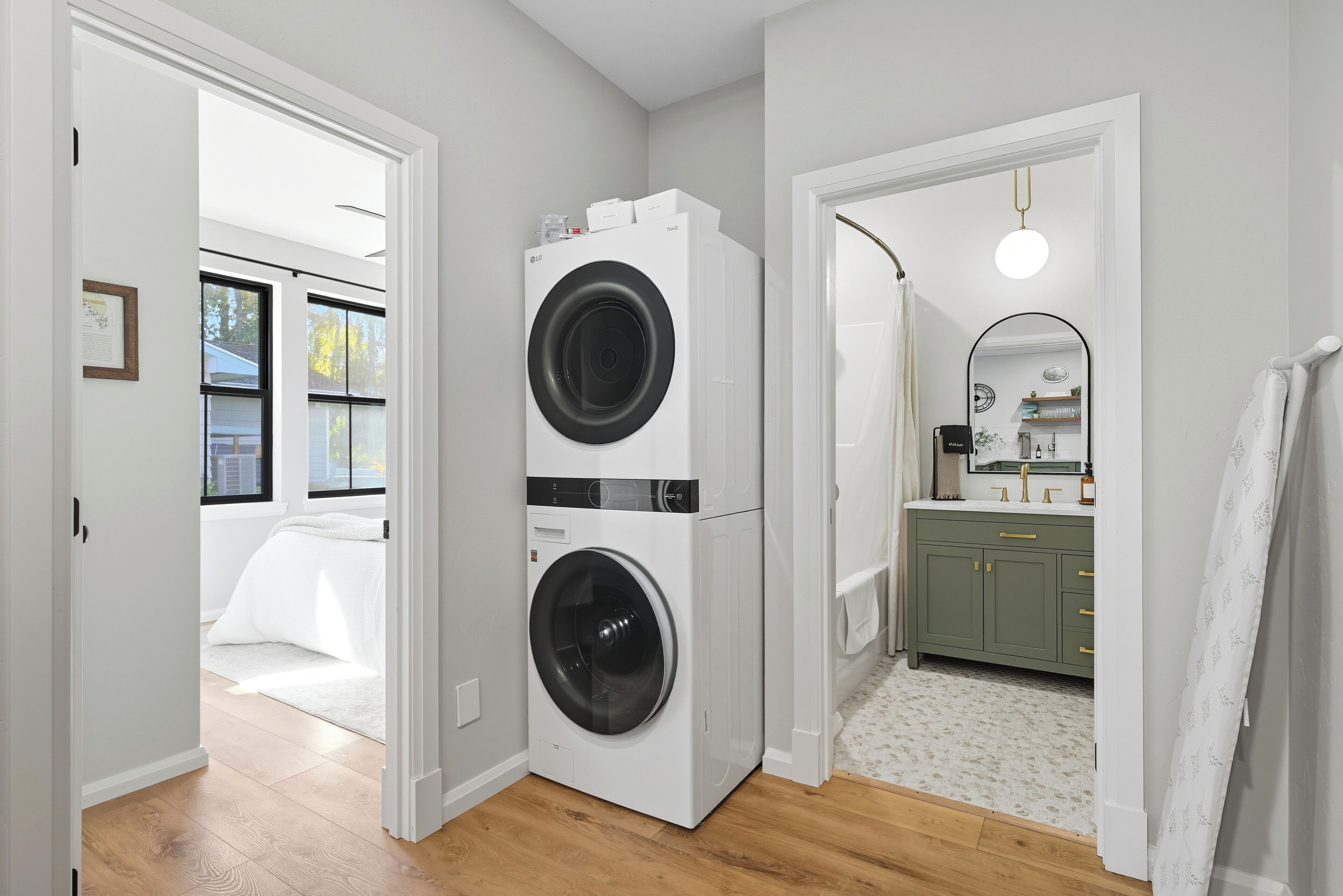 A laundry room with two stacked front-loading washing machines, a bathroom with a sink and mirror, and a bedroom with a bed and window.