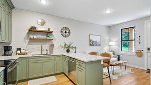 A modern kitchen with green cabinets, a marble countertop, and a dining area with a white rug and wooden chairs.