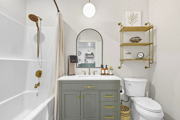 Un baño con bañera, inodoro, lavabo con espejo y repisa con objetos decorativos.