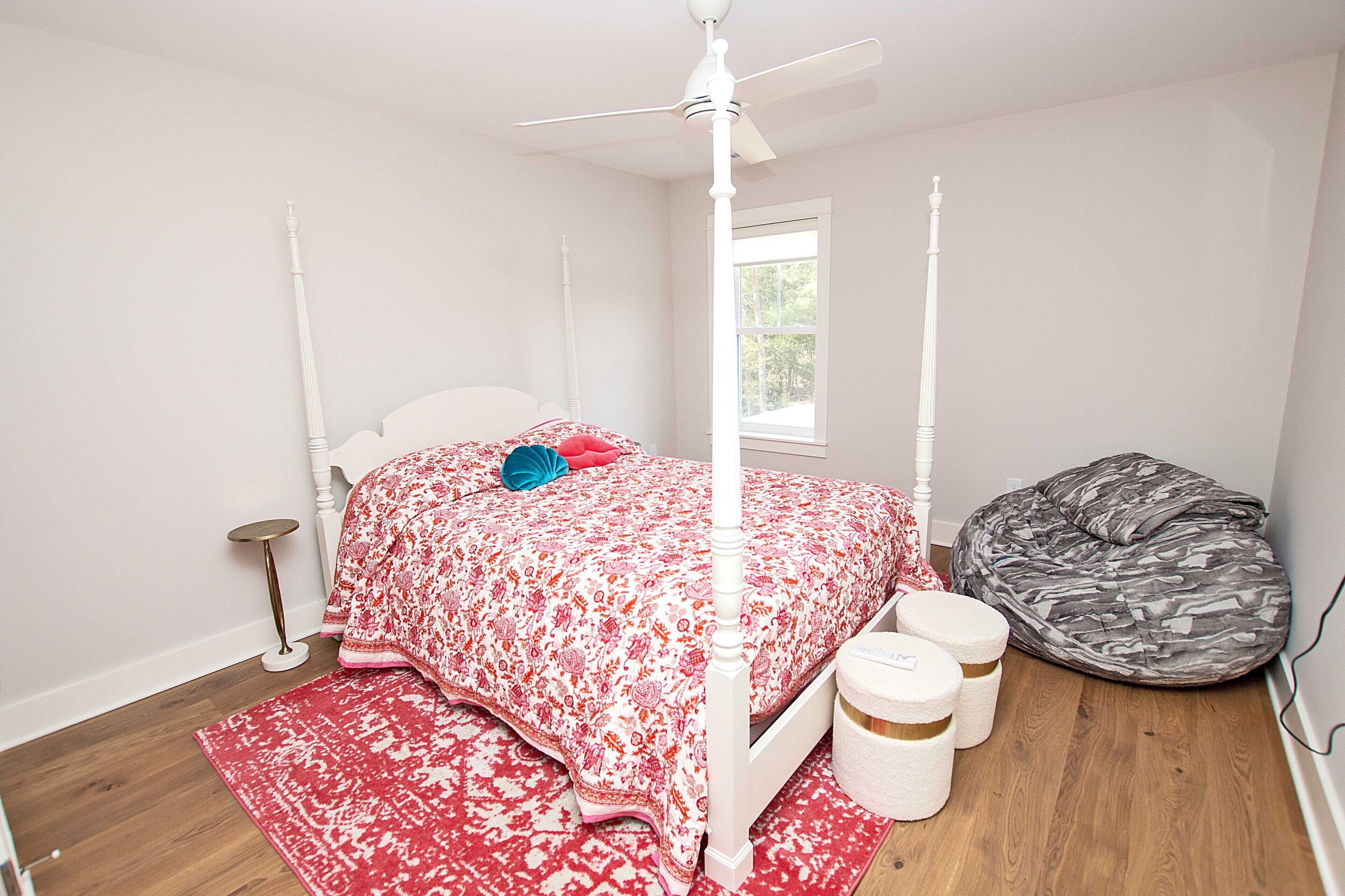 A bedroom with a four-poster bed, a ceiling fan, a window, a bean bag chair, and a small bedside table.
