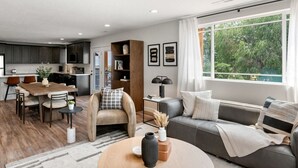 Une cuisine moderne avec des meubles de couleur sombre, un coin repas avec une table et des chaises en bois, un canapé gris et une grande fenêtre donnant sur un paysage verdoyant.