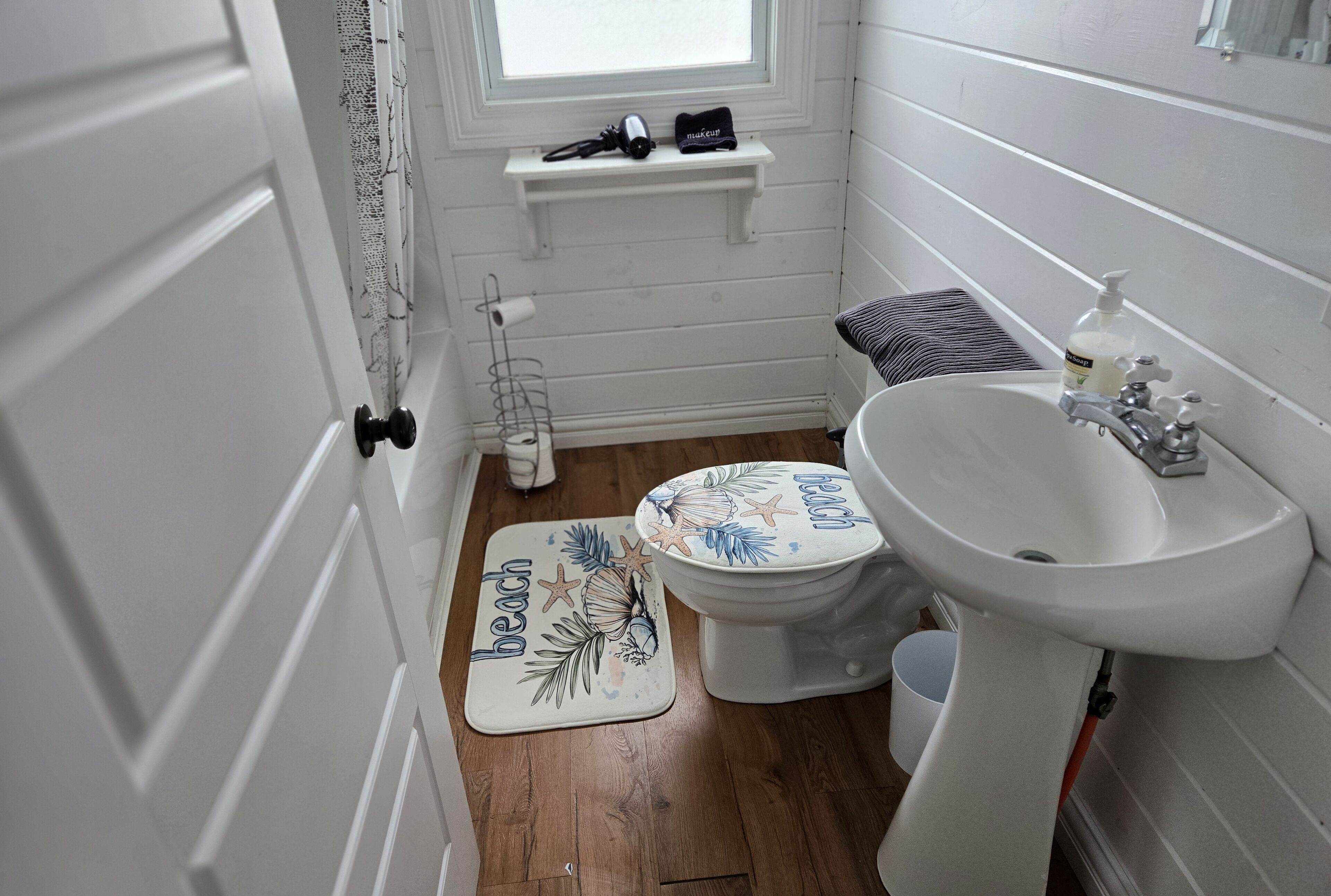 Une salle de bain avec un lavabo blanc, des toilettes et un rideau de douche. Il y a une étagère au-dessus des toilettes, sur laquelle se trouvent un sèche-cheveux et un sac.