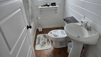 Une salle de bain avec un lavabo blanc, des toilettes et un rideau de douche. Il y a une étagère au-dessus des toilettes, sur laquelle se trouvent un sèche-cheveux et un sac.