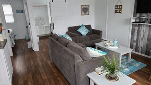 Een woonkamer met een hoekbank, een kleine tafel en een televisie. Er is een deur naar een andere kamer en een wandplank met decoratieve objecten.