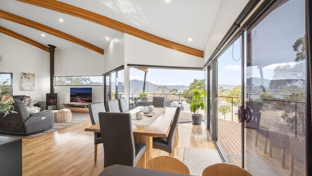 A modern living room with a wooden ceiling, a dining area, and large windows offering a view of the outdoors. - Tree Top Views - 4 Bedroom House (Rosetta)