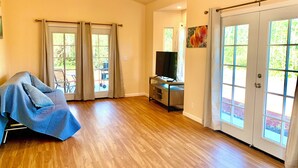 Un salon spacieux doté d’un plancher en bois, d’un canapé recouvert de tissu, d’un téléviseur posé sur un meuble et de grandes portes-fenêtres coulissantes donnant sur un espace extérieur.