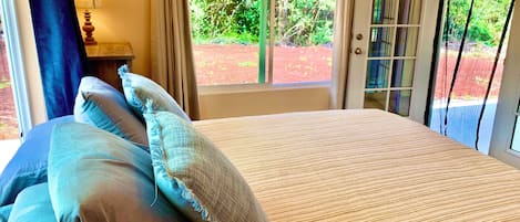A bedroom with a bed, pillows, a nightstand, a lamp, and a view of greenery through the window.